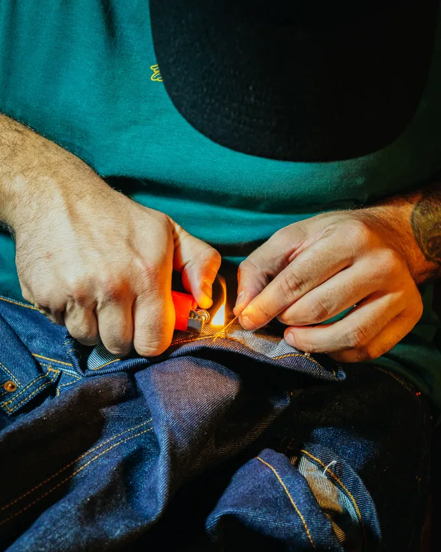 Dettaglio di mani che rifiniscono a mano un capo in denim con accendino e filo