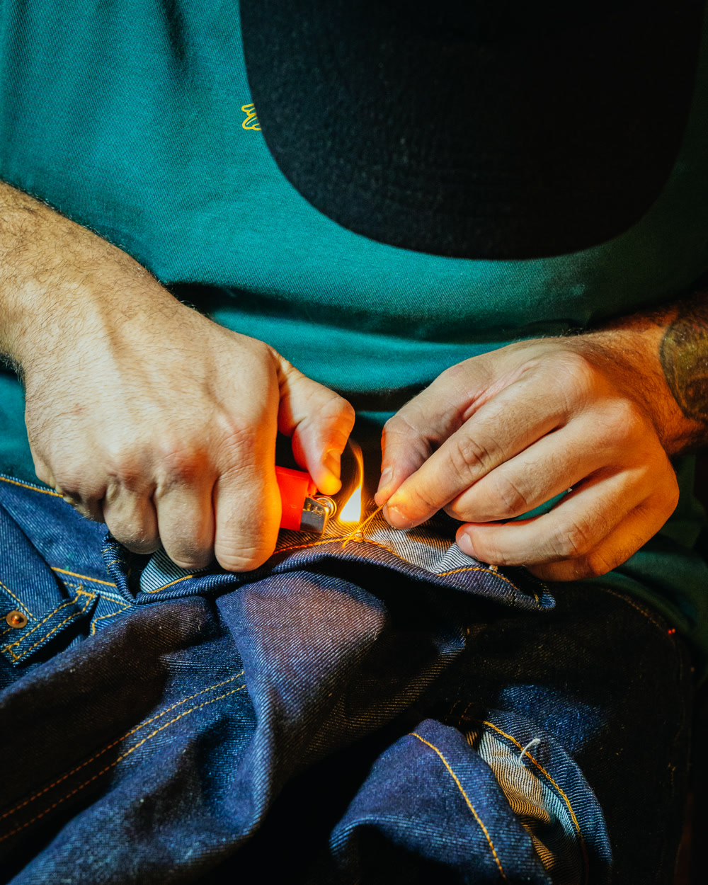 Dettaglio di mani che rifiniscono a mano un capo in denim con accendino e filo